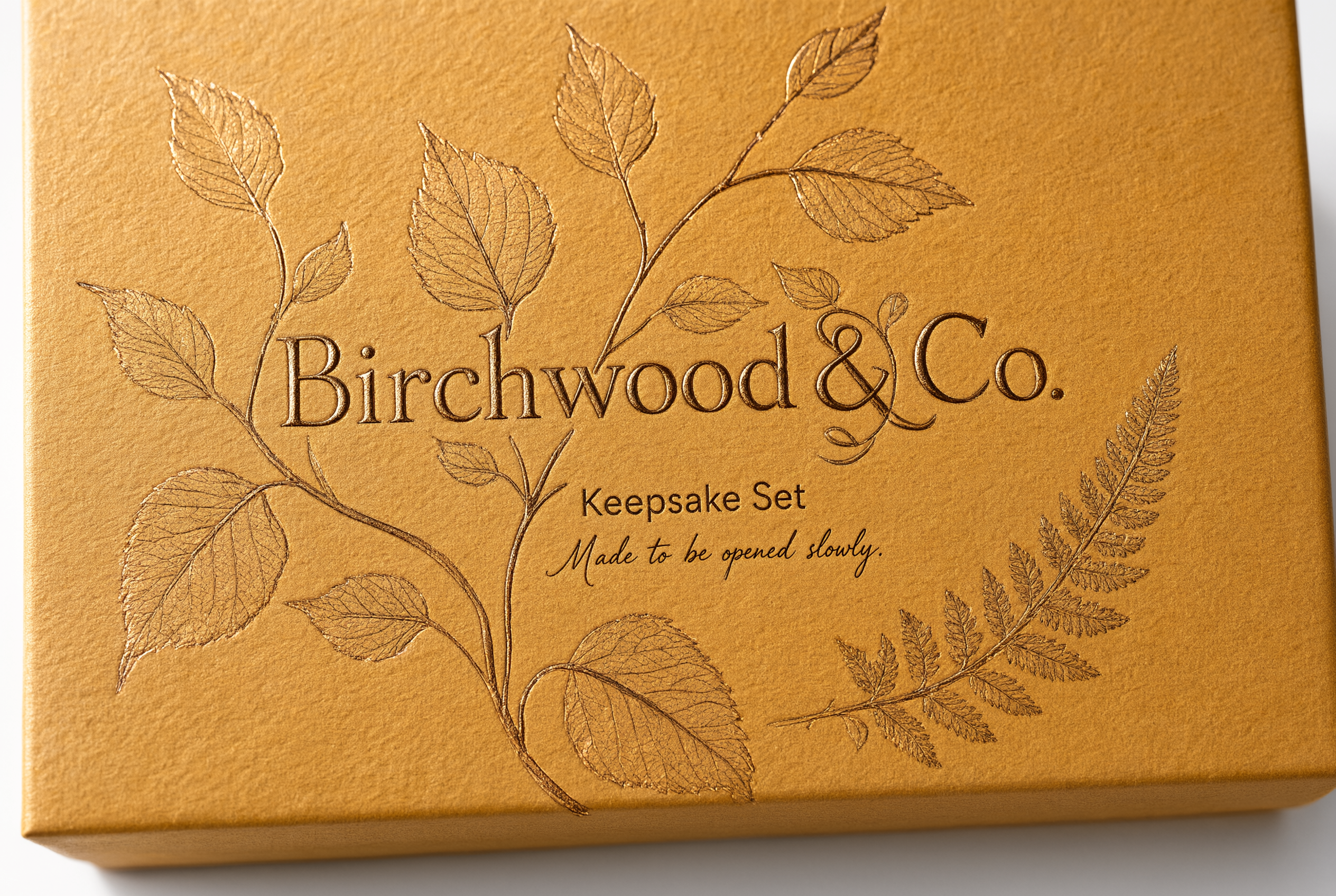 Close-up of bronze foil stamping on honey amber rigid paperboard showing the botanical motif