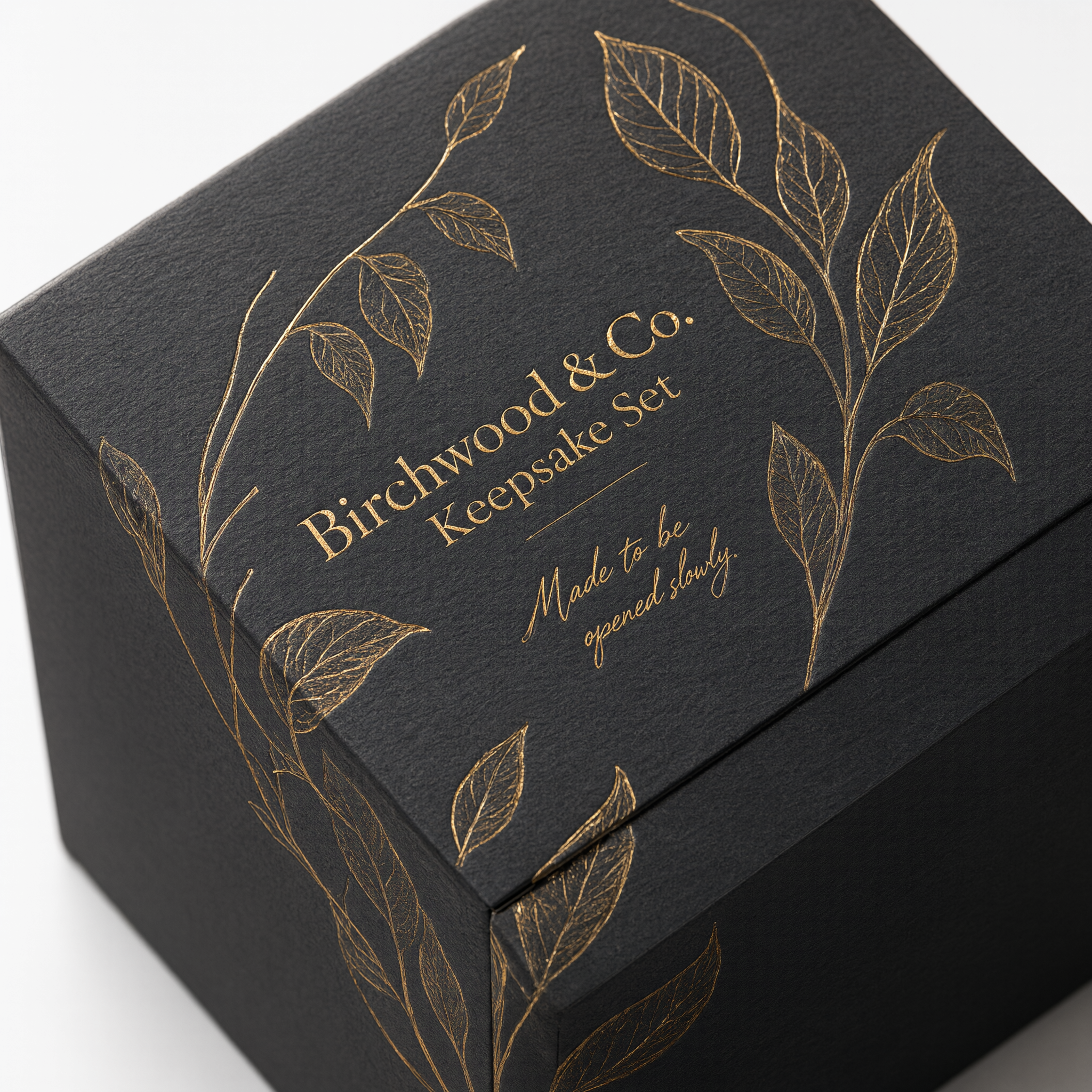 Close-up of gold foil stamping on charcoal rigid paperboard showing the botanical motif